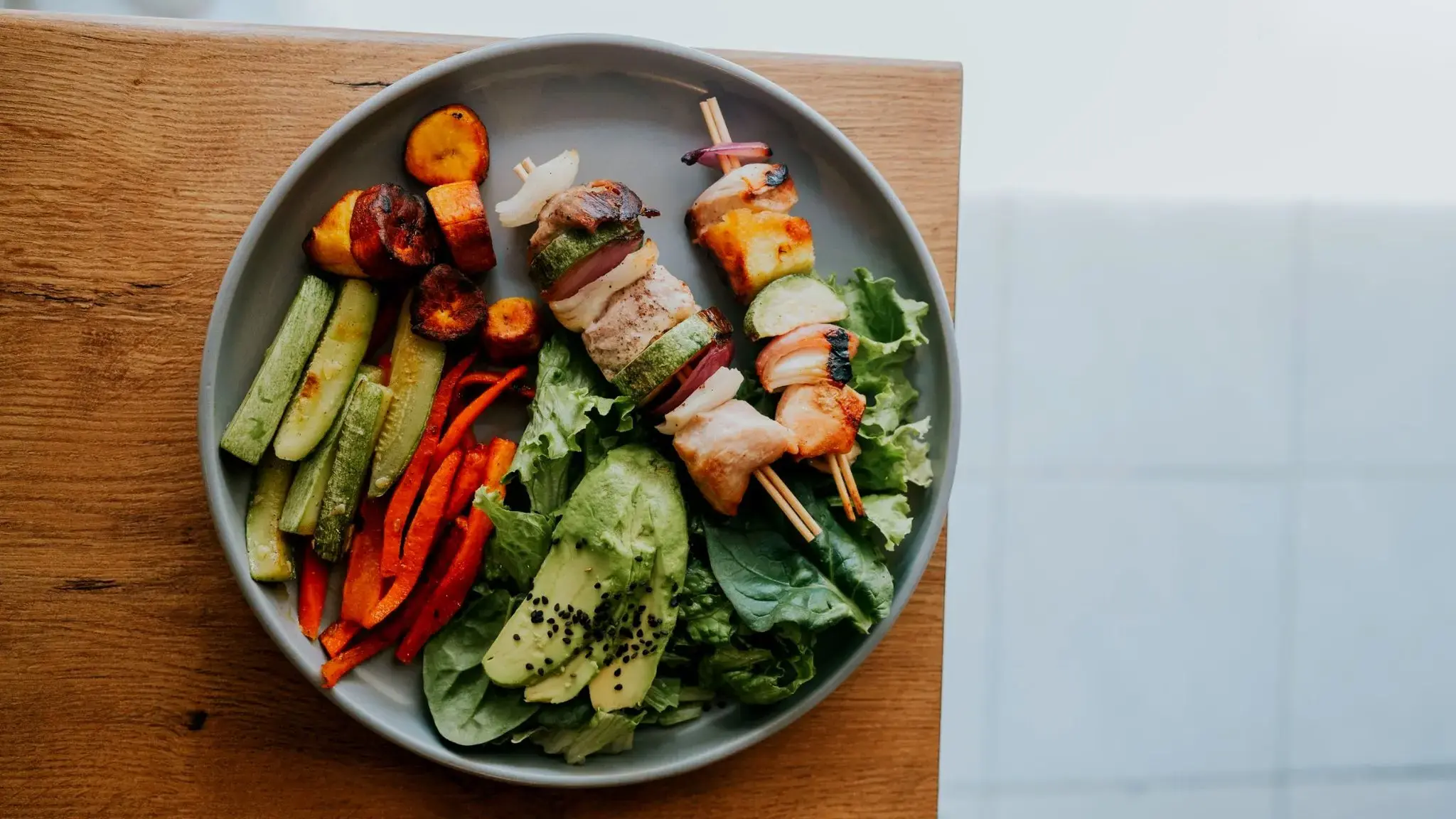 Ausgewogener Teller von oben: Blattsalat mit Avocado, Paprika- und Zucchinistreifen, Hähnchenspieße und geröstete Kochbananen auf einem Holzbrett